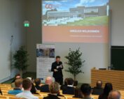 Ein Mann spricht in einem Hörsaal der Hochschule Coburg zum Publikum. Auf dem Bildschirm ist eine Präsentationsfolie mit dem Titel "Herzlich Willkommen" zu sehen. Auf der Folie ist ein modernes Gebäude abgebildet. Die Zuhörer sitzen auf Holzbänken, neben der Leinwand stehen zwei Topfpflanzen.