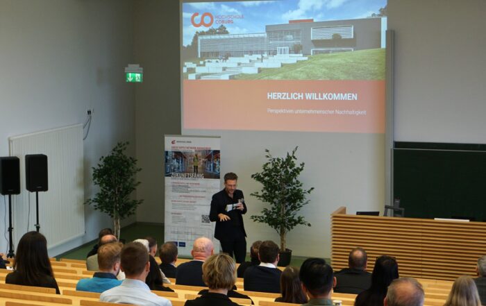 Ein Mann spricht in einem Hörsaal der Hochschule Coburg zum Publikum. Auf dem Bildschirm ist eine Präsentationsfolie mit dem Titel "Herzlich Willkommen" zu sehen. Auf der Folie ist ein modernes Gebäude abgebildet. Die Zuhörer sitzen auf Holzbänken, neben der Leinwand stehen zwei Topfpflanzen.