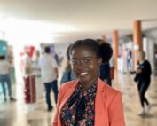 Eine Person lächelt in die Kamera, während sie in einem hellen, belebten Innenraum der Hochschule Coburg steht. Sie trägt eine Brille, ein geblümtes Hemd und einen orangefarbenen Blazer. Im unscharfen Hintergrund sind Menschen und Stände zu sehen.