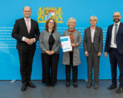 Fünf Personen in formeller Kleidung stehen vor einem blauen Hintergrund mit Text der Hochschule Coburg. Eine Person in der Mitte hält mit beiden Händen ein Dokument, über ihnen prangt prominent das Wappen-Emblem.