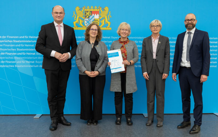 Fünf Personen in formeller Kleidung stehen vor einem blauen Hintergrund mit Text der Hochschule Coburg. Eine Person in der Mitte hält mit beiden Händen ein Dokument, über ihnen prangt prominent das Wappen-Emblem.
