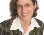 Eine Frau mit Brille, die einen grün-schwarz gestreiften Blazer über einem weißen Hemd trägt, lächelt in die Kamera und strahlt das Selbstbewusstsein einer Fachkraft der Hochschule Coburg aus. Der Hintergrund ist schlicht weiß.