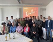 Eine Gruppe von Menschen hat sich in einem Raum versammelt und lächelt in die Kamera. Es scheint sich um eine Veranstaltung der Hochschule Coburg zu handeln. Im Hintergrund wird auf einer Projektionsfläche das Wort „Danke!“ angezeigt, während auf einem Tisch im Vordergrund einladende Getränke stehen, was eine feierliche und kameradschaftliche Atmosphäre schafft.