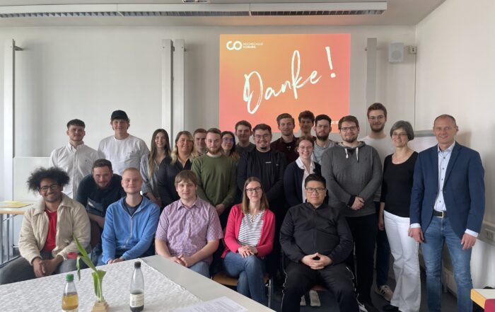 Eine Gruppe von Menschen hat sich in einem Raum versammelt und lächelt in die Kamera. Es scheint sich um eine Veranstaltung der Hochschule Coburg zu handeln. Im Hintergrund wird auf einer Projektionsfläche das Wort „Danke!“ angezeigt, während auf einem Tisch im Vordergrund einladende Getränke stehen, was eine feierliche und kameradschaftliche Atmosphäre schafft.