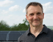 Ein Mann in einem dunklen Poloshirt steht im Freien und lächelt stolz vor den Solarmodulen auf dem Campus der Hochschule Coburg. Der Hintergrund zeigt einen teilweise bewölkten Himmel und üppiges Grün, was sowohl Innovation als auch Natur einfängt.