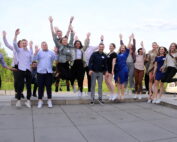 Eine Gruppe von Menschen in legerer Business-Kleidung springt im Gleichschritt auf einer gepflasterten Fläche mit malerischer Aussicht. Sie wirken fröhlich, während sie in der Nähe der Hochschule Coburg feiern. In der Ferne runden ein Wohnmobil und malerische Hügel die lebhafte Szene ab.