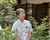 In einer üppigen Gartenlandschaft der Hochschule Coburg spricht eine Person mit Brille und weißem Hemd angeregt und gestikuliert mit den Händen. Grüne Pflanzen und eine Steinlaterne schaffen eine ruhige Kulisse.