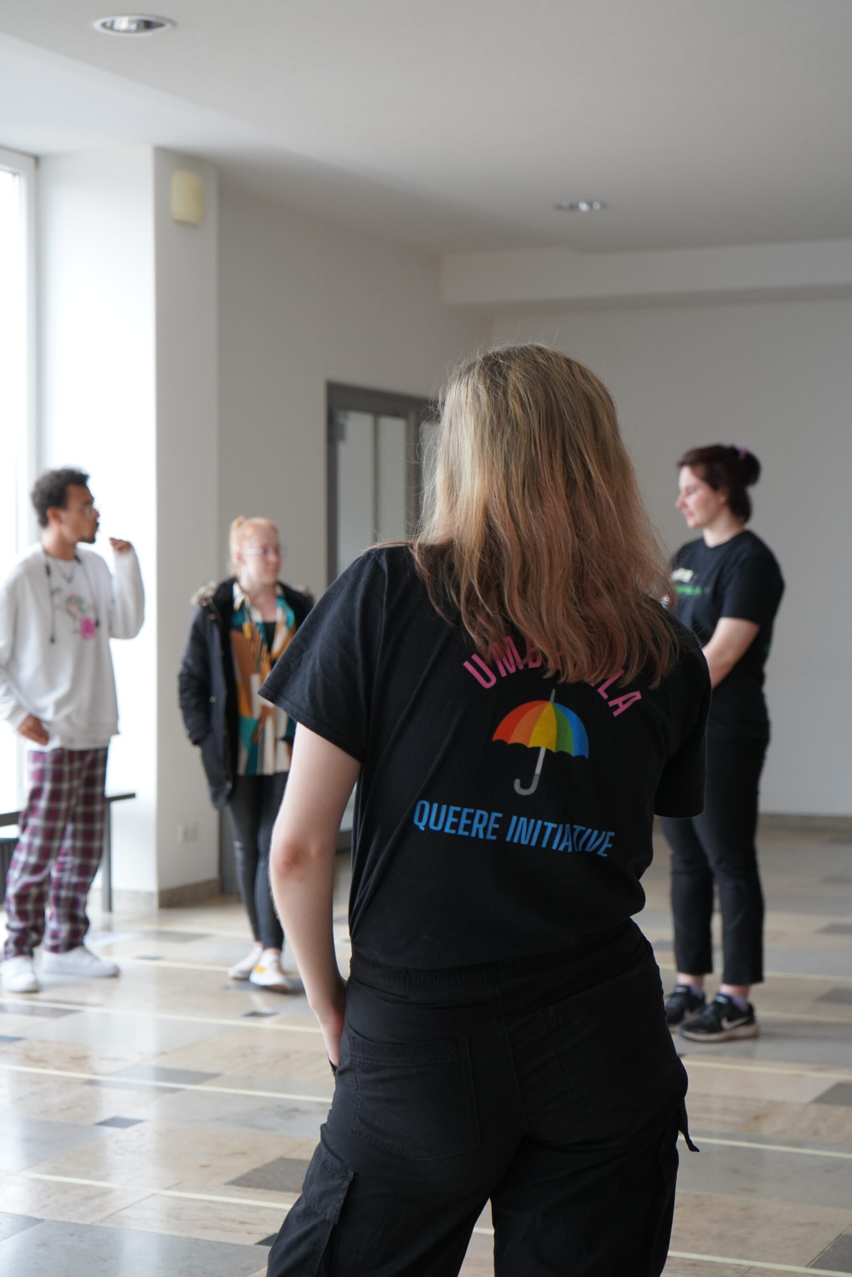 Eine Gruppe von vier Personen steht in einem Raum mit großen Fenstern der Hochschule Coburg. Eine Person im Vordergrund trägt ein schwarzes Shirt mit einem bunten Regenschirm und dem Text „UMs Ganze Queere Initiative“. Die anderen stehen sich gegenüber und unterhalten sich.