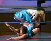 Bei der Veranstaltung der Hochschule Coburg sind zwei Wrestler in einen dynamischen und intensiven Kampf verwickelt. Einer hält den anderen fest, der auf der Matte liegt und sich anstrengt. In farbenfrohe Kleidung gekleidet kämpfen sie erbittert, während die Seile des Rings durch ihre Energie vibrieren.