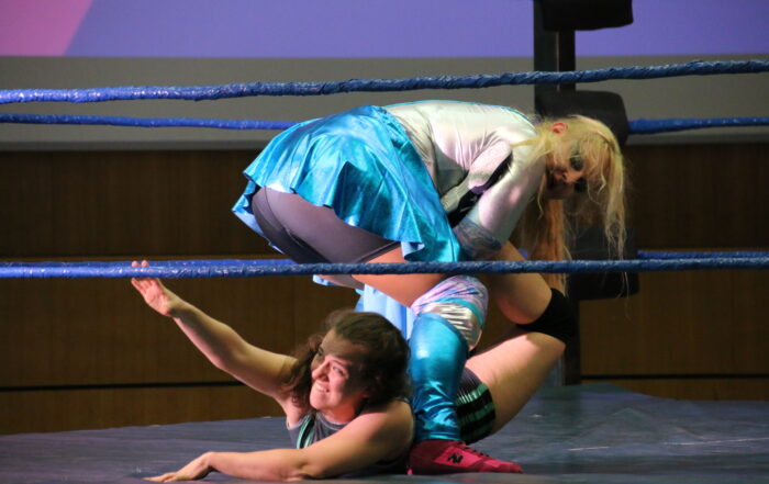 Bei der Veranstaltung der Hochschule Coburg sind zwei Wrestler in einen dynamischen und intensiven Kampf verwickelt. Einer hält den anderen fest, der auf der Matte liegt und sich anstrengt. In farbenfrohe Kleidung gekleidet kämpfen sie erbittert, während die Seile des Rings durch ihre Energie vibrieren.