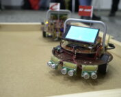 Beim Wettbewerb der Hochschule Coburg stehen zwei kleine Roboter auf einer flachen Holzfläche. Jeder Roboter verfügt über einen Bildschirm, auf dem Daten angezeigt werden, sowie über verschiedene Sensoren und Kabel. Aus ihren Rücken ragen anmutig kleine Flaggen hervor, die zur dynamischen Atmosphäre der Ausstellung beitragen.