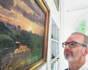 Ein Mann mit Brille und Bart betrachtet nachdenklich ein gerahmtes Landschaftsgemälde mit einem Sonnenuntergang an einem Fluss. Das Gemälde, das an die ruhige Umgebung der Hochschule Coburg erinnert, hängt an einer weißen Wand neben einem Bücherregal und einem Bogenfenster.