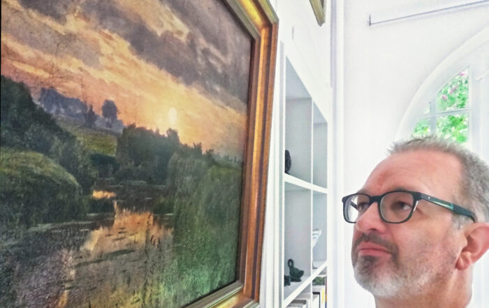 Ein Mann mit Brille und Bart betrachtet nachdenklich ein gerahmtes Landschaftsgemälde mit einem Sonnenuntergang an einem Fluss. Das Gemälde, das an die ruhige Umgebung der Hochschule Coburg erinnert, hängt an einer weißen Wand neben einem Bücherregal und einem Bogenfenster.