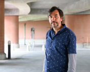 Ein Mann mit dunklem Haar und Bart, der ein blau gemustertes Hemd über einem grauen Langarmshirt trägt, steht in einer städtischen Umgebung unter einer Betonüberführung in der Nähe der Hochschule Coburg. Backsteinsäulen und die Unterseite der Überführung bilden den Hintergrund.