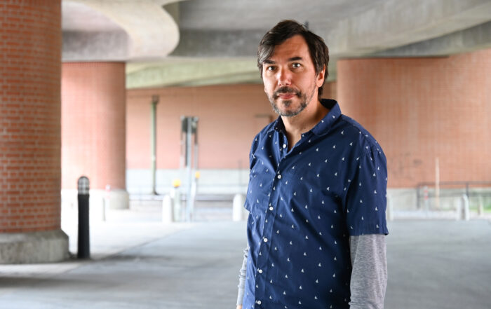Ein Mann mit dunklem Haar und Bart, der ein blau gemustertes Hemd über einem grauen Langarmshirt trägt, steht in einer städtischen Umgebung unter einer Betonüberführung in der Nähe der Hochschule Coburg. Backsteinsäulen und die Unterseite der Überführung bilden den Hintergrund.