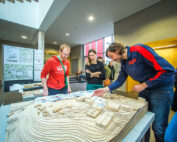 Drei Personen betrachten ein großes, detailreiches Architekturmodell einer Landschaft mit Gebäuden. Im Hintergrund zeigen Schautafeln Diagramme und Pläne der Hochschule Coburg. Die Kulisse scheint ein überdachter Ausstellungsraum mit Tageslicht zu sein.