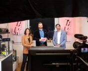 Drei Personen stehen hinter einem Schreibtisch mit Mikrofonen in einem Studioaufbau der Hochschule Coburg und lächeln in die Kamera. Im Hintergrund ist in fetten Buchstaben das Wort „TALK“ zu lesen, im Vordergrund ist eine Kamera zu sehen.