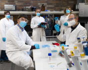Fünf Wissenschaftler der Hochschule Coburg führen in Schutzkleidung Experimente im Labor durch. Sie tragen weiße Laborkittel, blaue Handschuhe und Gesichtsmasken, während sie mit verschiedenen Geräten arbeiten und Notizen machen. Das Labor besteht aus einer Ziegelwand und Regalen voller wissenschaftlicher Instrumente.