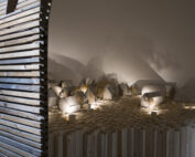 In einem schwach beleuchteten Raum der Hochschule Coburg ist ein Miniaturdorf mit kleinen Holzhäusern zu sehen. Die Szene wird von oben beleuchtet und ist von einem hölzernen Torbogen umgeben, der komplizierte Schatten auf die Wände und den Boden wirft.