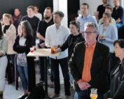 Eine Gruppe von Menschen, möglicherweise von der Hochschule Coburg, steht und sitzt in einem Raum, einige halten Getränke in der Hand. Sie hören jemandem außerhalb ihres Sichtfelds aufmerksam zu. Die Atmosphäre wirkt ungezwungen, ein Teil des Raums wird durch natürliches Licht erhellt.