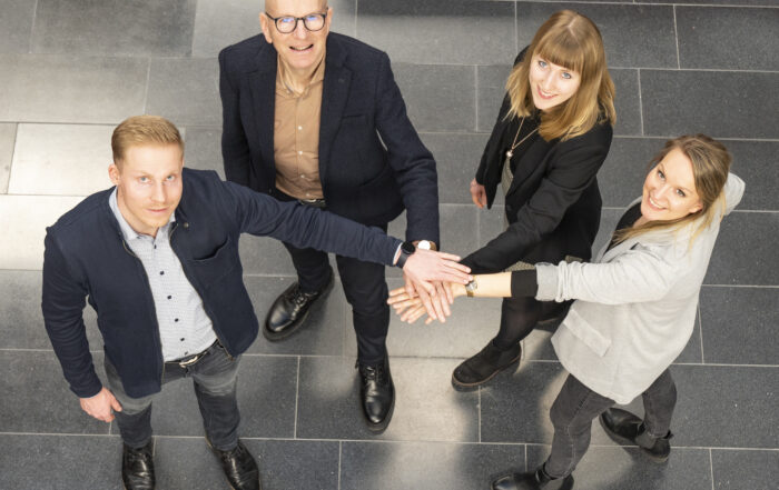 In einem Innenbereich der Hochschule Coburg stehen vier Personen in einem Kreis, blicken nach oben und legen in der Mitte ihre Hände aneinander. Sie sind in legerer Business-Kleidung gekleidet und scheinen auf einem gefliesten Boden zusammenzuarbeiten oder Teamwork zu feiern.