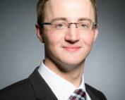 Ein Mann mit kurzen braunen Haaren und Brille lächelt. Er trägt einen schwarzen Anzug, ein weißes Hemd und eine karierte Krawatte und verkörpert damit den professionellen Geist der Hochschule Coburg. Der Hintergrund ist mit einem schlichten Farbverlauf versehen.