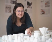 Eine Person mit Brille steht lächelnd neben einem Tisch, der mit geometrischen weißen Tonmodellen bedeckt ist, an der Hochschule Coburg. Im Hintergrund hängen gedruckte Informationsblätter an der Wand.