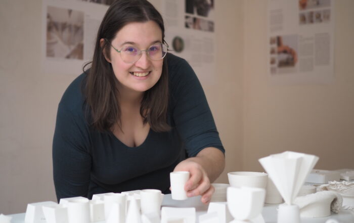 Eine Person mit Brille steht lächelnd neben einem Tisch, der mit geometrischen weißen Tonmodellen bedeckt ist, an der Hochschule Coburg. Im Hintergrund hängen gedruckte Informationsblätter an der Wand.