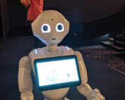 Ein humanoider Roboter mit einem Bildschirm auf der Brust zeigt Text und Zahlen an. Er hat große runde Augen und leuchtende Lichter auf den Schultern. Er steht auf einem Teppich in einem Gebäude der Hochschule Coburg. Im Hintergrund ist eine Person in einem rosa Hemd zu sehen.