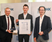Drei Männer stehen in formeller Kleidung zusammen. Der Mann links hält eine Medaille und ein Zertifikat der Hochschule Coburg, der Mann in der Mitte hält ein großes gerahmtes Zertifikat und der Mann rechts lächelt. Sie stehen vor einem Hintergrund mit dem Text „IHK“.
