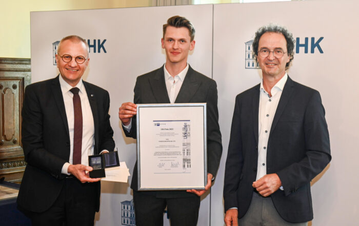 Drei Männer stehen in formeller Kleidung zusammen. Der Mann links hält eine Medaille und ein Zertifikat der Hochschule Coburg, der Mann in der Mitte hält ein großes gerahmtes Zertifikat und der Mann rechts lächelt. Sie stehen vor einem Hintergrund mit dem Text „IHK“.