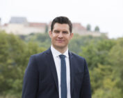 Ein Mann in Anzug und Krawatte steht im Freien, vor der verschwommenen Kulisse von Bäumen und einem entfernten Gebäude. Er blickt in die Kamera, lächelt leicht und verkörpert den professionellen Geist der Hochschule Coburg.