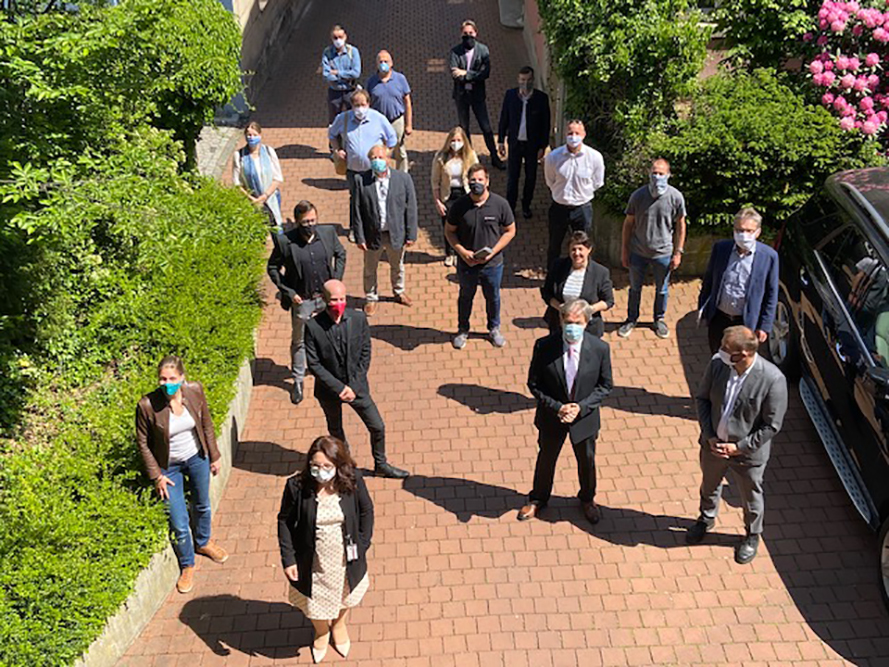 Eine Gruppe maskierter Menschen steht draußen auf einem Ziegelweg an der Hochschule Coburg, umgeben von üppigem Grün. Sie stehen mit etwas Abstand zueinander und blicken unter einem sonnigen Himmel in die Kamera.