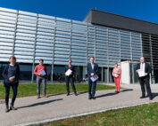 Eine Gruppe von sechs Personen steht in einer Reihe vor dem modernen Gebäude der Hochschule Coburg mit großen Fenstern und glatten Metallpaneelen. Sie halten Ordner in den Händen und wahren unter dem klaren blauen Himmel Abstand zueinander.