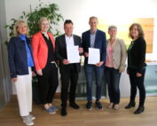 Sechs Personen stehen in einem Gebäude, das wie ein Büro der Hochschule Coburg aussieht, und lächeln in die Kamera. In der Mitte halten zwei Männer Dokumente in der Hand, umgeben von drei Frauen und einem weiteren Mann. Eine Pflanze verleiht der professionellen Umgebung einen Hauch von Grün.