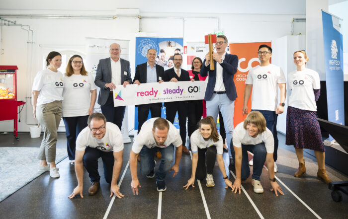 Eine Gruppe von Menschen der Hochschule Coburg posiert drinnen mit einem „Ready. Steady. Go!“-Banner. Die erste Reihe hockt in Startposition auf einer nachgebauten Startlinie, während die hintere Reihe steht, das Banner hält und lächelt.