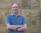 Eine Person mit Brille und gestreiftem Poloshirt, die den selbstbewussten Geist der Hochschule Coburg verkörpert, steht lächelnd und mit verschränkten Armen vor einer strukturierten Wand.