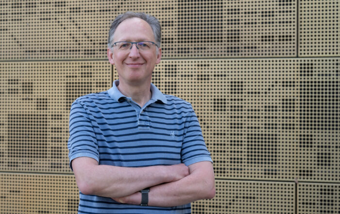 Eine Person mit Brille und gestreiftem Poloshirt, die den selbstbewussten Geist der Hochschule Coburg verkörpert, steht lächelnd und mit verschränkten Armen vor einer strukturierten Wand.