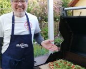 Ein Mann mit Brille, weißem Hemd und dunkelblauer Schürze steht lächelnd neben einem Grill. Er zeigt auf eine Pizza mit Rucola und Tomaten in dieser bezaubernden Außenkulisse der Hochschule Coburg, eingerahmt von üppigem Grün und einem Holzzaun.