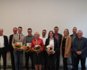 Eine Gruppe von elf Personen steht in einer Reihe, einige halten Blumensträuße und Zertifikate in den Händen und feiern eine Leistung an der Hochschule Coburg. Die Szene ist in einem Innenbereich vor einer schlichten weißen Wand. Alle sind in Business- oder Abendgarderobe gekleidet und lächeln in die Kamera.