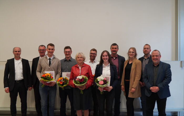 Eine Gruppe von elf Personen steht in einer Reihe, einige halten Blumensträuße und Zertifikate in den Händen und feiern eine Leistung an der Hochschule Coburg. Die Szene ist in einem Innenbereich vor einer schlichten weißen Wand. Alle sind in Business- oder Abendgarderobe gekleidet und lächeln in die Kamera.
