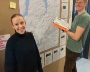 Zwei Personen stehen neben einer großen Karte, die an einem schwarzen Brett der Hochschule Coburg befestigt ist. Die rechte Person hält ein offenes Buch und lächelt, während die linke Person ebenfalls in die Kamera lächelt.
