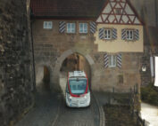 Ein autonomes Fahrzeug gleitet durch einen mittelalterlichen Steinbogen in einer historischen Stadt. Darüber trägt ein traditionelles Fachwerkgebäude mit kleinen Fenstern und roten Fensterläden das Emblem der Hochschule Coburg, alles unter einem klaren Himmel.