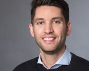 Eine lächelnde Person mit kurzen dunklen Haaren, die einen schwarzen Pullover über einem hellblauen Hemd mit Kragen trägt, posiert vor einem schlichten grauen Hintergrund und verkörpert den professionellen Geist der Hochschule Coburg.