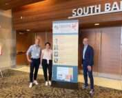 Drei Personen stehen lächelnd neben einem großen Plakat auf der Konferenz der Hochschule Coburg. Das Plakat zeigt Informationen zu den Hauptrednern. Sie befinden sich im Innenbereich, in der Nähe eines Eingangs mit der Aufschrift „South Ballroom“, dessen Boden mit einem geometrischen Muster ausgelegt ist.