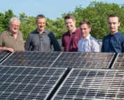 Fünf Männer der Hochschule Coburg stehen gemeinsam lächelnd hinter Solarmodulen inmitten des üppigen Grüns und unter einem strahlend blauen Himmel.