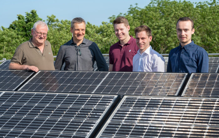 Fünf Männer der Hochschule Coburg stehen gemeinsam lächelnd hinter Solarmodulen inmitten des üppigen Grüns und unter einem strahlend blauen Himmel.