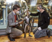 Zwei Menschen mit Masken und Handschuhen knien neben einer kleinen Rüstung in einem Museum der Hochschule Coburg. Eine Person richtet den Helm, während die andere die Rüstung stützt. Im Hintergrund sind weitere Rüstungen ausgestellt.