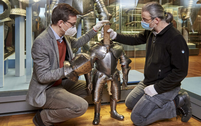 Zwei Menschen mit Masken und Handschuhen knien neben einer kleinen Rüstung in einem Museum der Hochschule Coburg. Eine Person richtet den Helm, während die andere die Rüstung stützt. Im Hintergrund sind weitere Rüstungen ausgestellt.