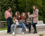 Fünf Frauen sitzen mit Laptops und Notizbüchern auf einer Bank im Freien an der Hochschule Coburg. Sie lächeln und unterhalten sich vor der üppig grünen Kulisse eines Parks.
